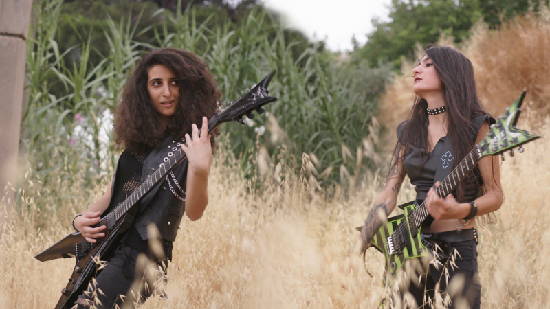 Lilas Mayassi and Shery Bechara stand in a field wearing all-black holding their guitars.