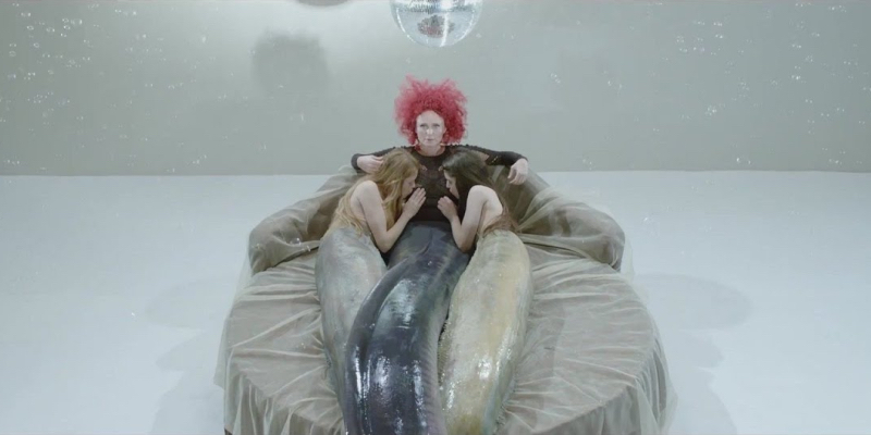 Two young women with long fish tails suck the nipples of an older woman with red hair who also has a long fish body.