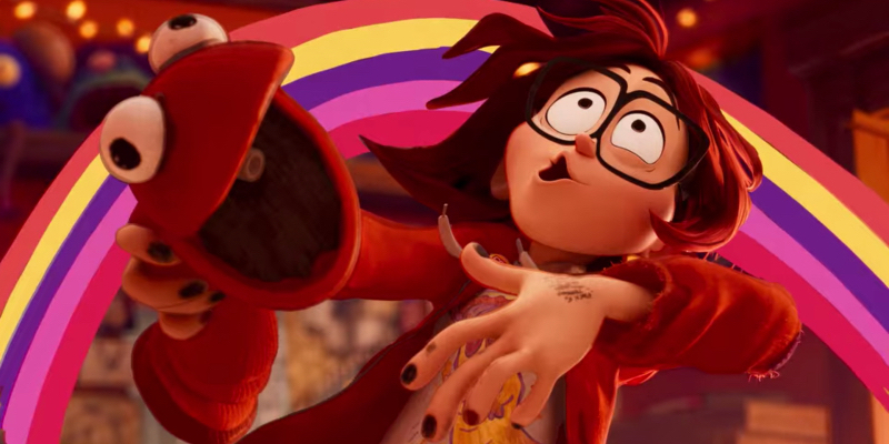 An animated teen girl with glasses and black nail polish leans back in front of a rainbow while wearing a red puppet.