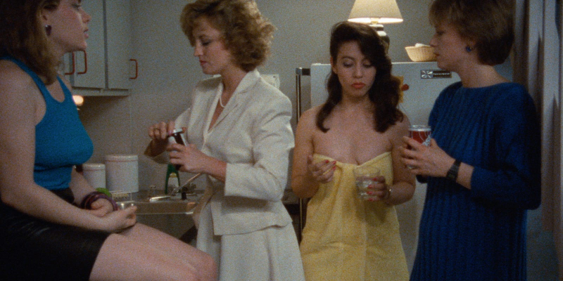 Four women squeeze into a tight kitchen. Two are wearing professional attire, one is wearing a tank top, and one just has on a towel. 