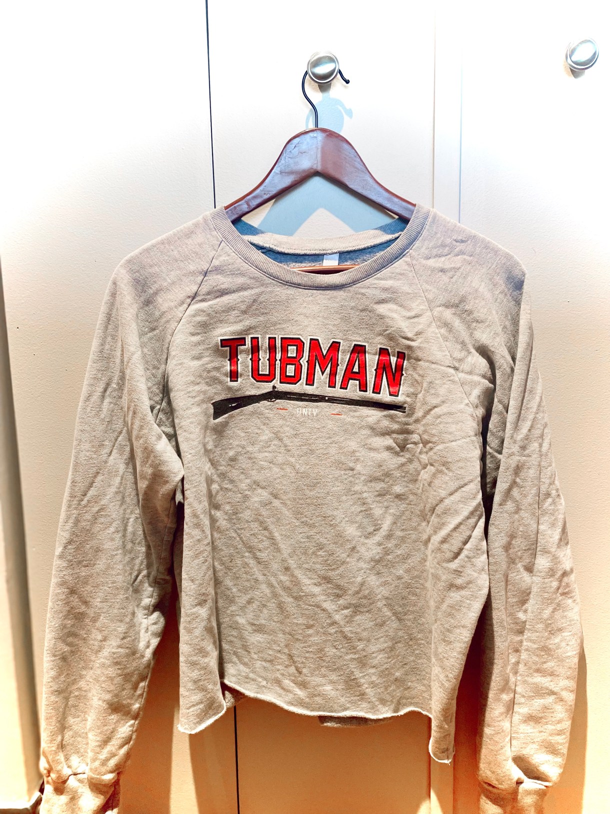 A wrinkled grey sweatshirt that says "Tubman University" in black and red lettering over an insignia of a rifle. It's on a hanger against a wall at sunset, which is giving everything a gold hue.