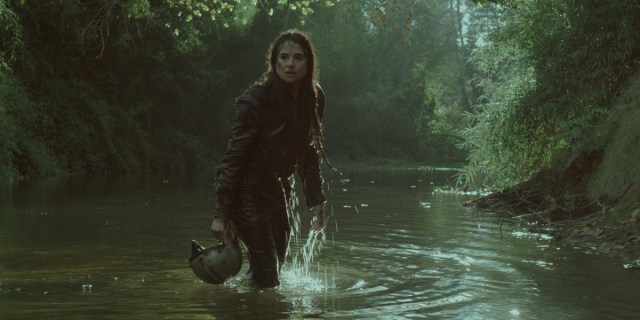 A woman emerges from a lush green river holding a motorcycle helmet.
