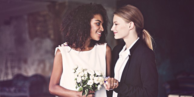 A Black women in a white dresss holding flowers looks lovingily at a white woman in a suit on their wedding day