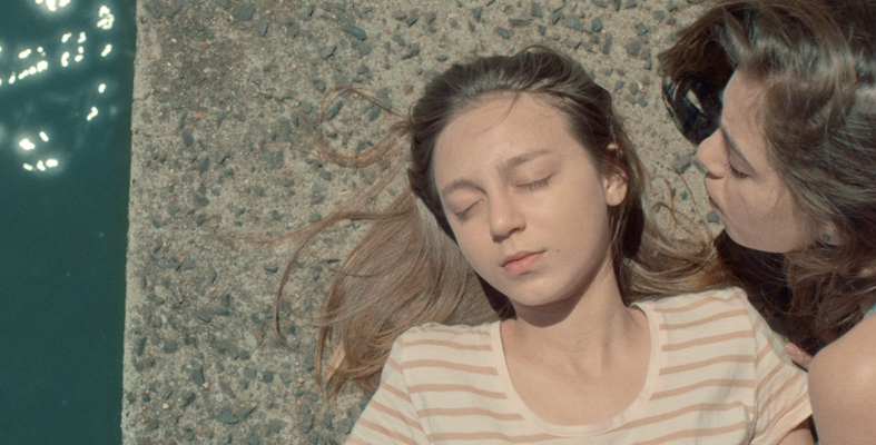 A young girl lies next to a body of water with her eyes closed as another young girl hovers over her blowing on her hair.