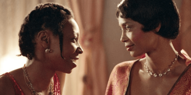 Celie and Shug, both Black brown skinned women, sit on a bed and look lovingly into each others eyes.