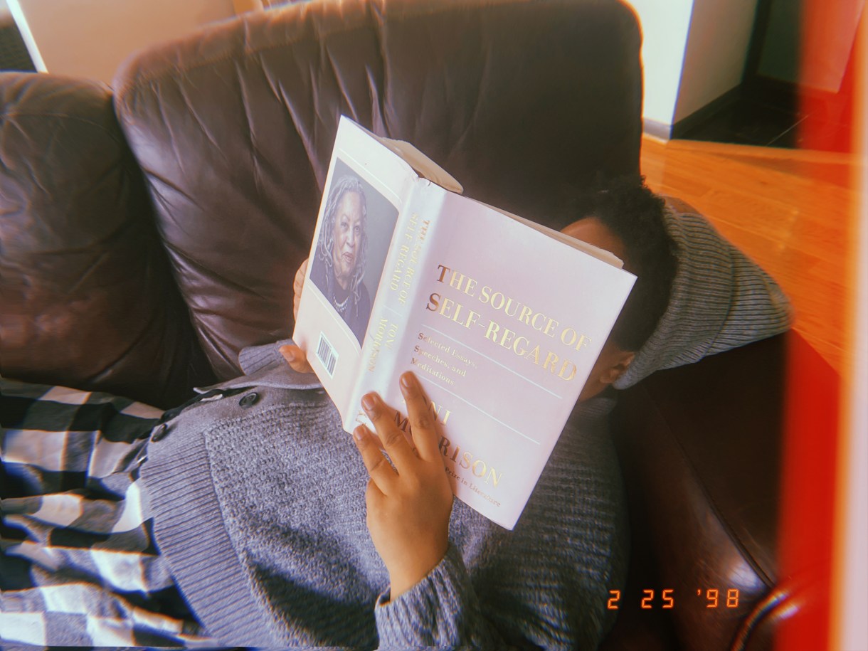 Carmen reads The Source of Self-Regard by Toni Morrison while lying on her couch. Carmen is a Black woman who is wearing a gray sweater and matching gray beanie. Carmen's face is behind the book in this photo.