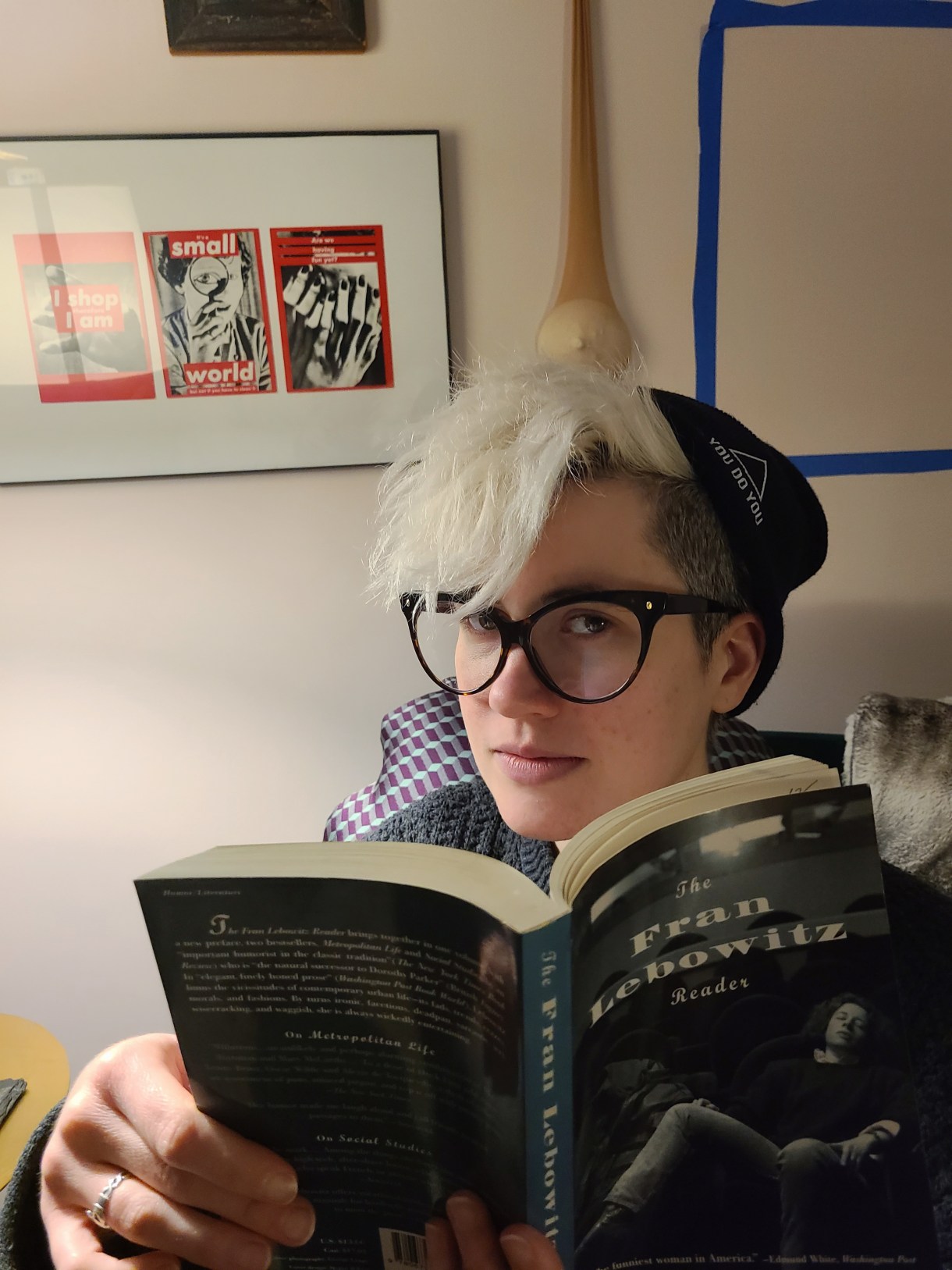 Nicole looks up from reading The Fran Lebowitz Reader. Nicole is a white genderqueer human wearing a black beanie, with bleached hair and glasses.