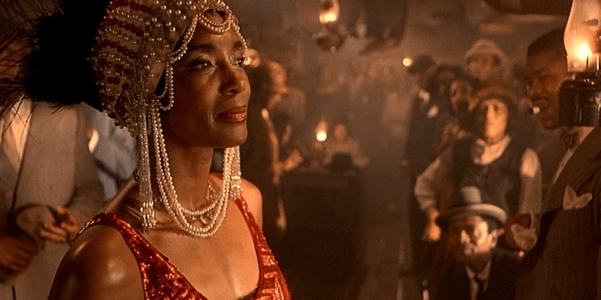 Shug stands on stage, decked out in a pearl headpiece, with her head held high as others look at her ready to perform.