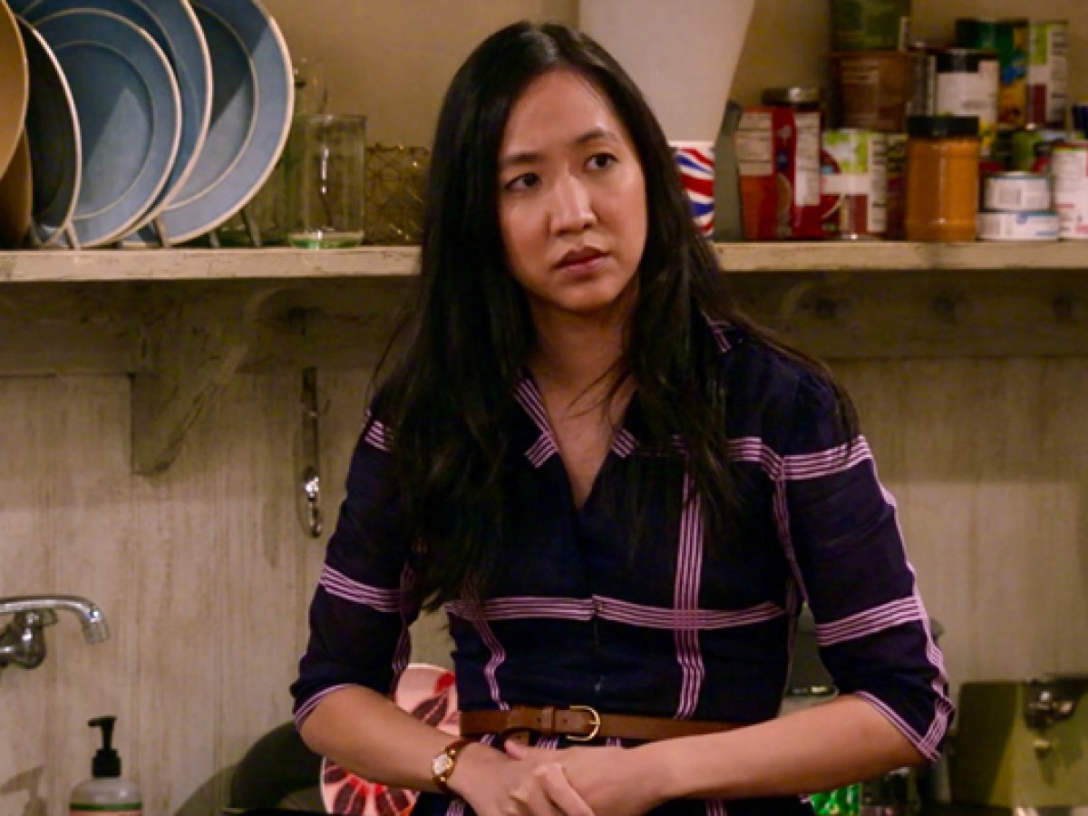 Ellen in How I Met Your Father, an Asian lesbian, is in a flannel purple dress and a confused look on her face in front of a kitchen wall with pots hanging above her head.