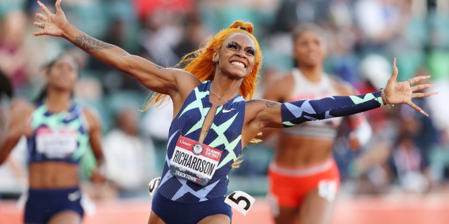 Sha'Carri Richardson with orange hair and long nails celebrates crossing the finish line during a race.