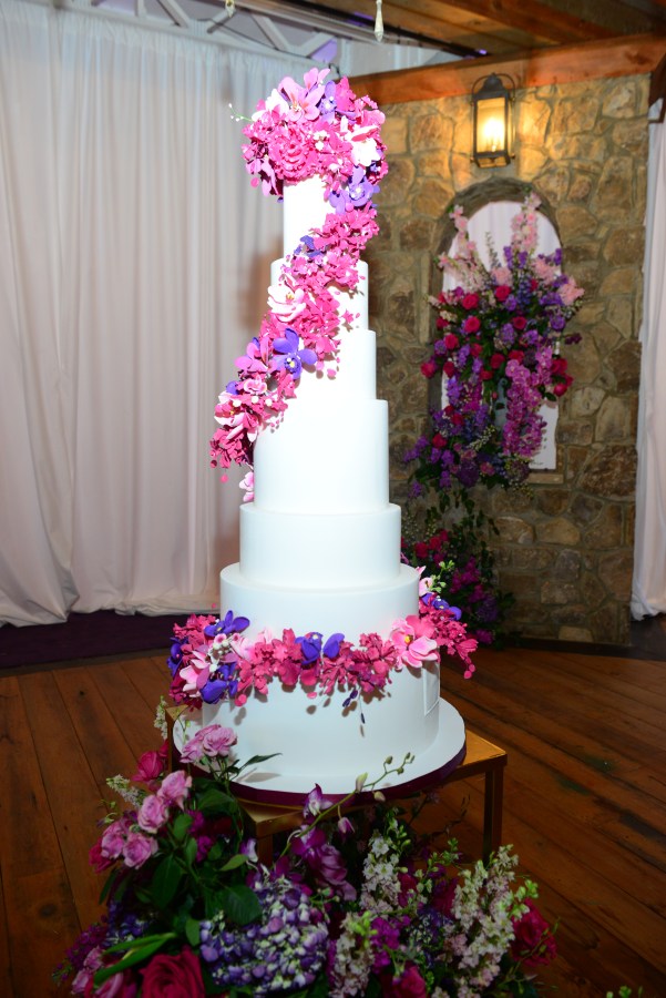 A seven tier white wedding cake with pink and purple flowers cascading down the side.