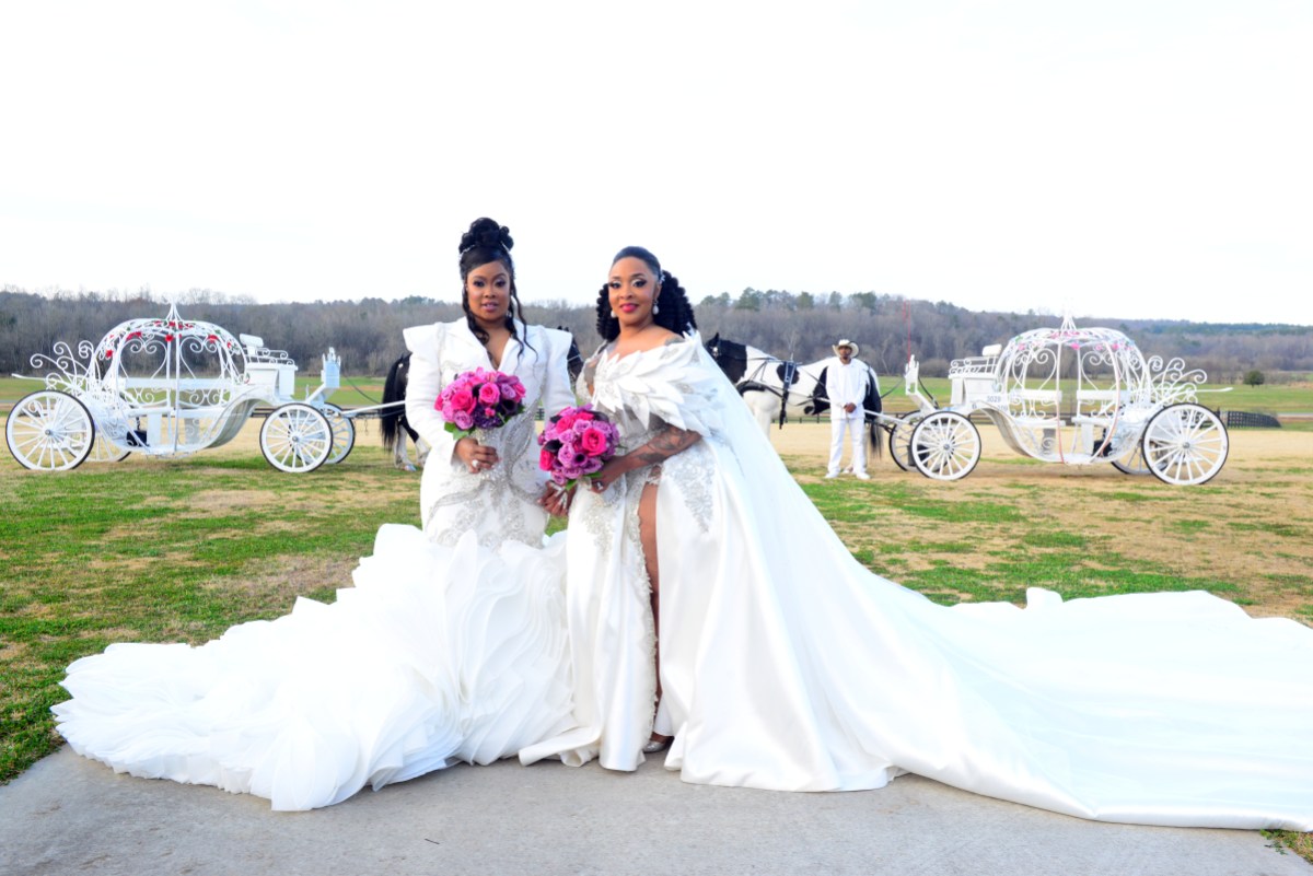 Da Brat & Jesseca “Judy” Dupart pose in matching gowns and up do's for their wedding, they are outside holding pink and purple wedding bouquets. Behind them on the grass of the estate are two white carriages that look like they are from Cinderella.
