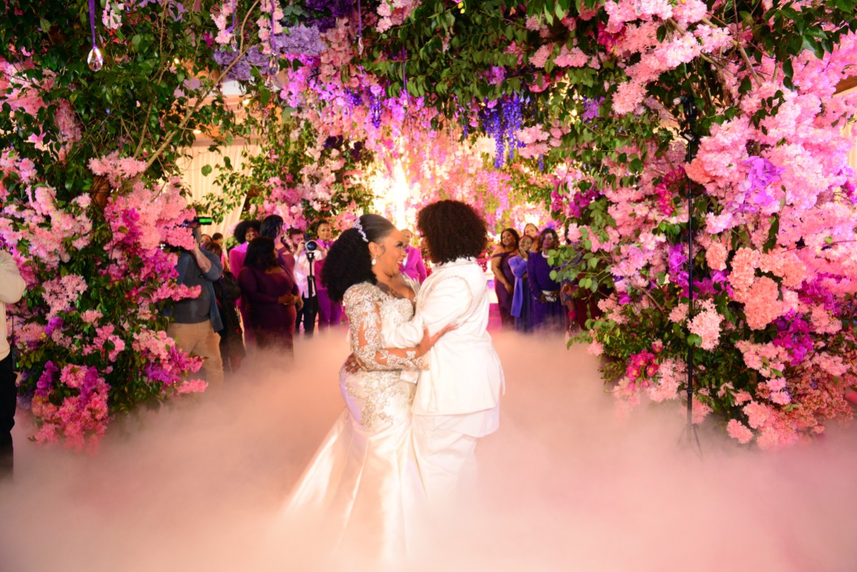 Da Brat & Jesseca “Judy” Dupart hold each other while dancing at their wedding, Da Brat is in a white suit with curls in a bob hair cut and Judy is in a white mermaid gown. There are purple and pink flowers everywhere and romantic mist beneath their feet.