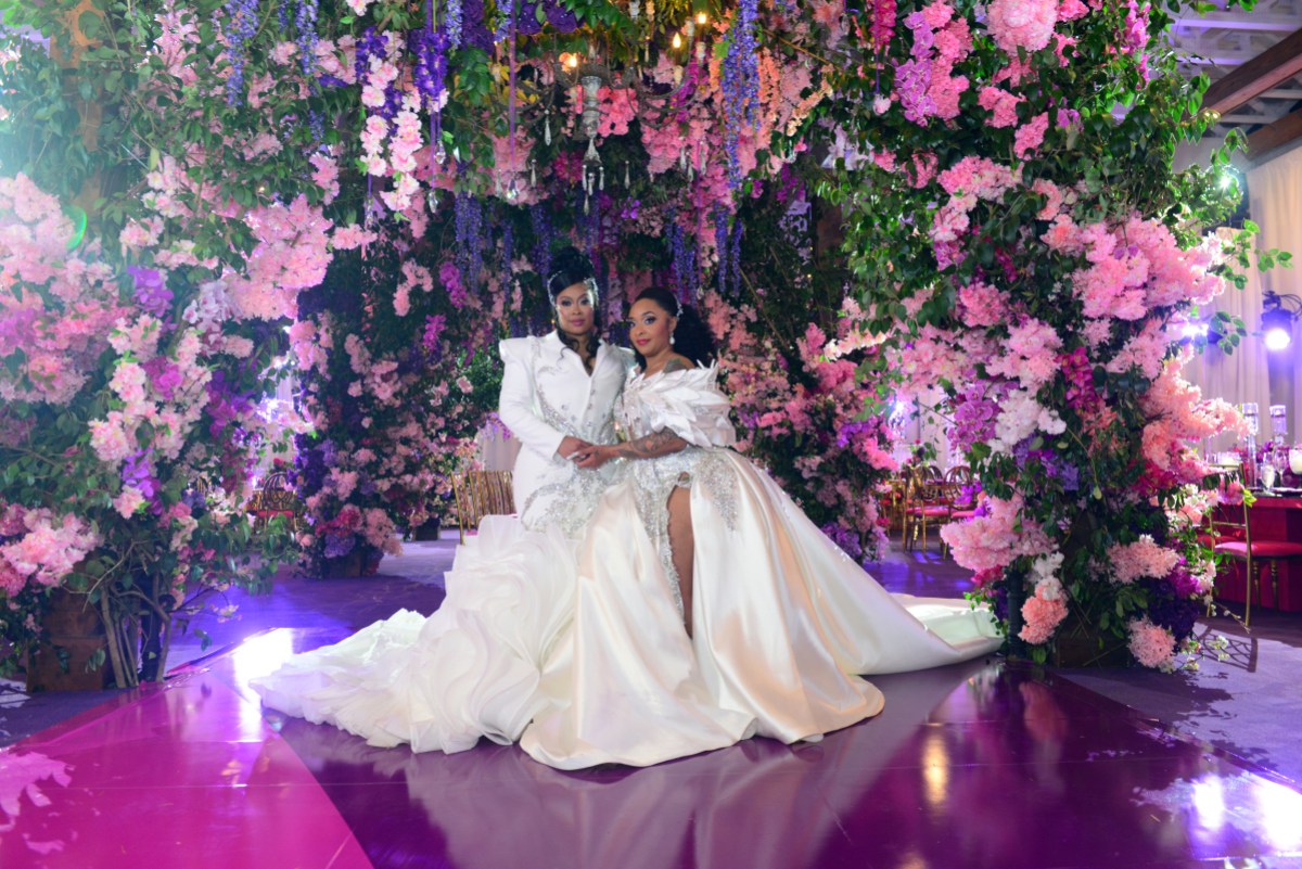 Da Brat & Jesseca “Judy” Dupart hold each other and pose for a wedding photo in matching white gowns and up-do's. They have purple and pink flowers.