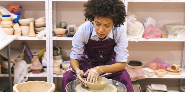 a Black woman leans over a pottery wheel upon which she appears to be making a clay dish. Her hands are covered in clay, and she is wearing a pair of purple coveralls over a blue collared shirt.