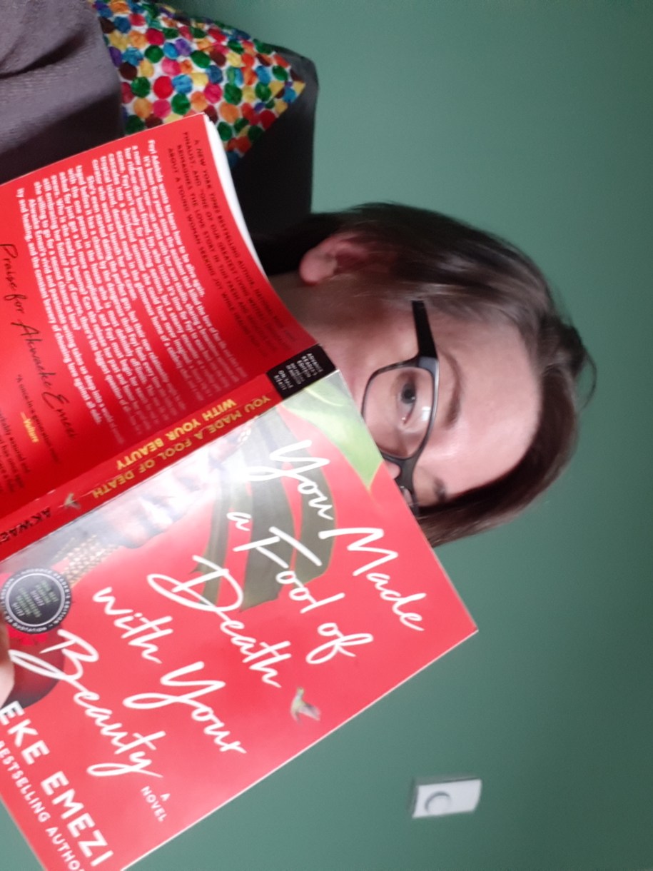 Casey looks over the top of a book with one eye. The book is "You Made a Fool of Death With Your Beauty." Casey is a white woman wearing a sweater with black glasses