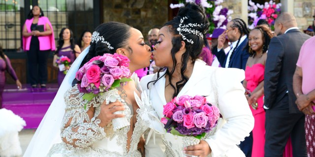 Da Brat & Jesseca “Judy” Dupart kiss at their wedding. There is a minister in a black pantsuit and pink coat behind them, and an arch of purple flowers. Wedding guess look on and smile, all of them wearing shades of pink and purple. MICHAEL SEABROOK/WE TV