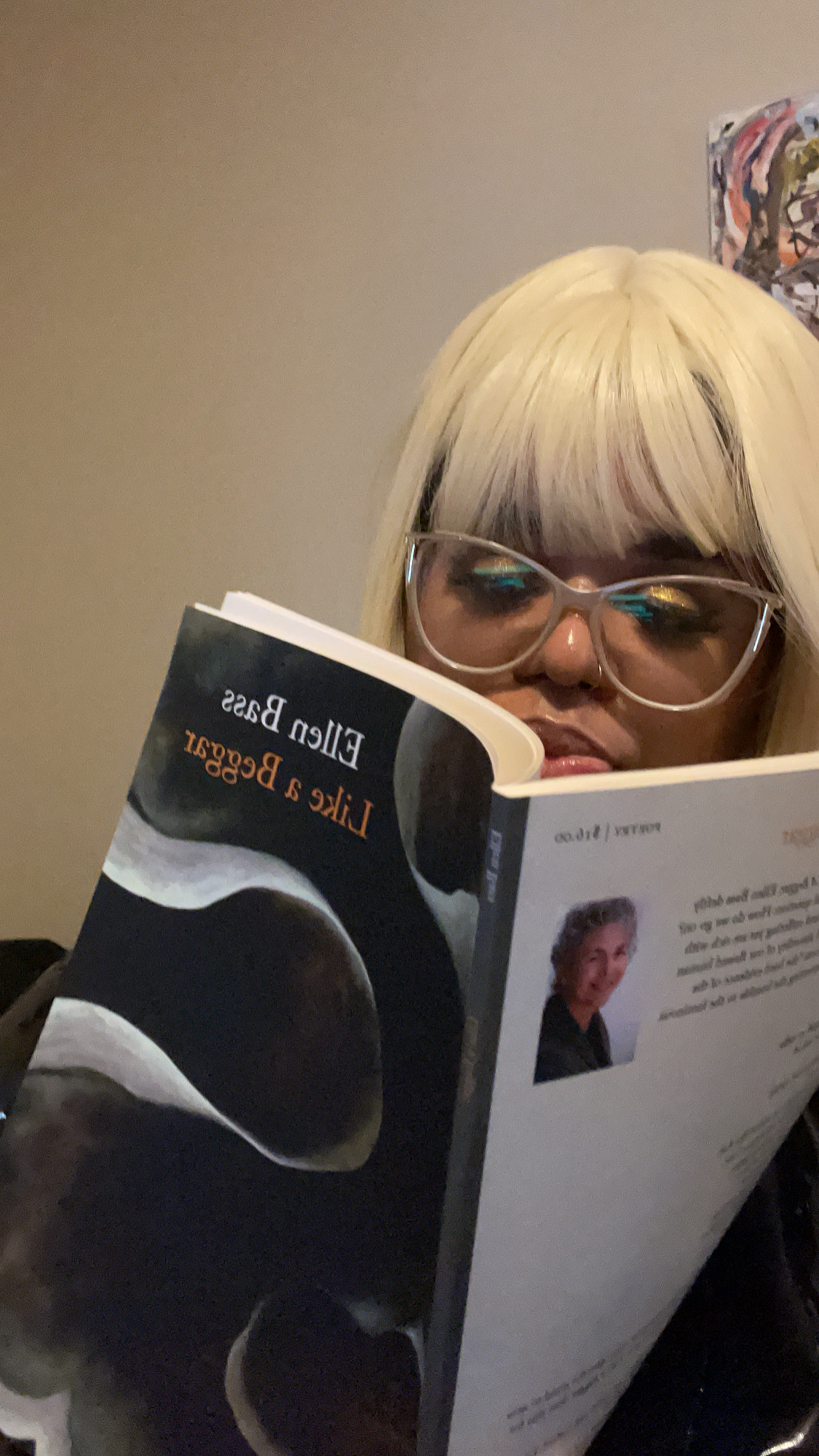 Dani reads from "Like a Beggar" by Ellen Bass. Dani is a Black woman wearing clear glasses and a platinum wig. You can just see her face and hair popping up behind the book.