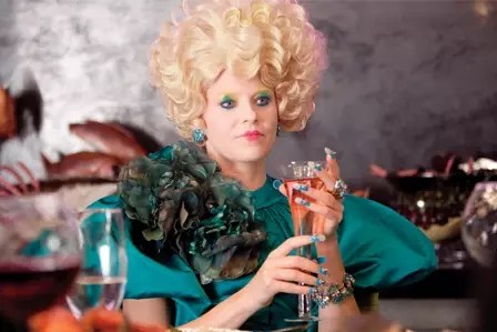 an image of Effie Trinket from the hunger games wearing an extremely opulent teal outfit with eye makeup and fancy nails and holding some kind of alcoholic beverage