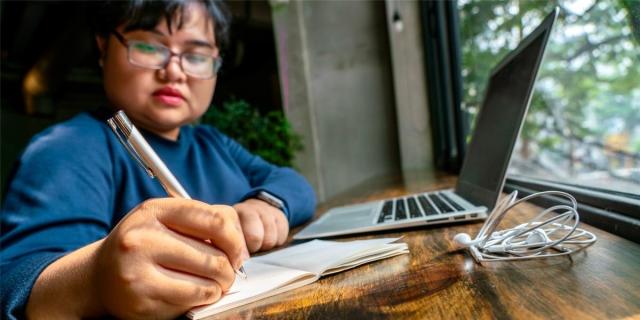 An Asian person with short hair and pink lipstick is writing in a notebook next to a MacBook computer, at a wooden desk overlooking a window with greenery.
