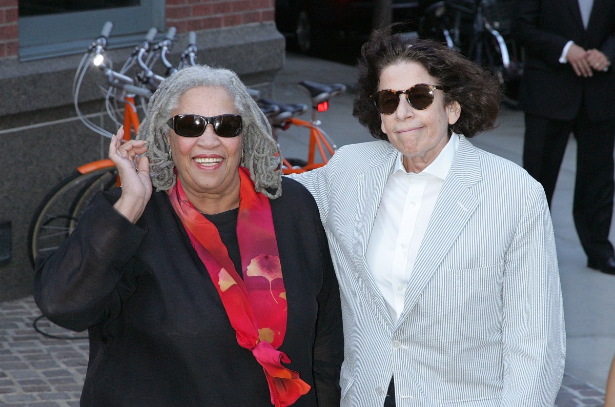 Toni Morrison is in a black dress with a red scarf and sunglasses, Fran Lebowitz is also in sunglasses with a grey suit coat and her arm around Toni.