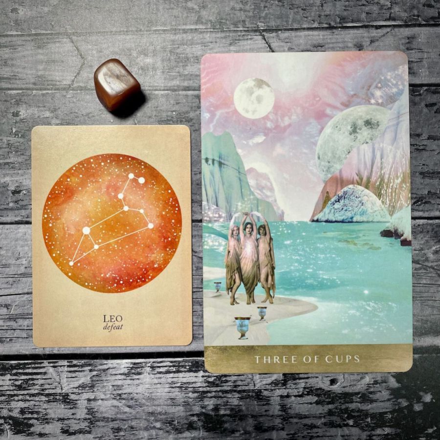 A card says Leo: defeat with an orange circle, next to a card that says three of cups with an illustration of three women in flapper dresses next to a moonlit lake a sunset