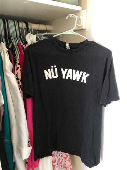 A black tee shirt with the words NÜ YAWK in white on it. It is hanging in a closet.