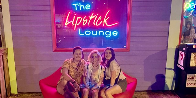 The three hosts of the podcast "Cruising," two queer women and a non-binary person, sit on a couch shaped like lips in from of a sign that says "The Lipstick Lounge" in neon