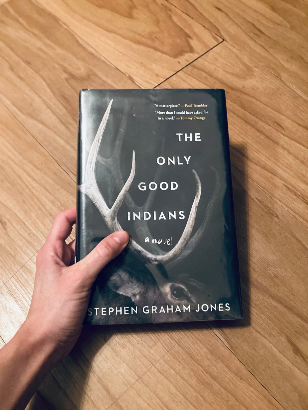 Ro holds a book in frame. The book is "The Only Good Indians" by Stephen Graham Jones. Ro is a white person and their hand is white.