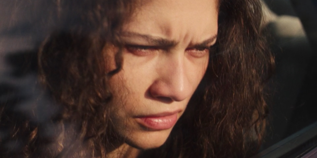 A close up on Zendaya as Rue through a car window. Her eyes are red and she looks upset.
