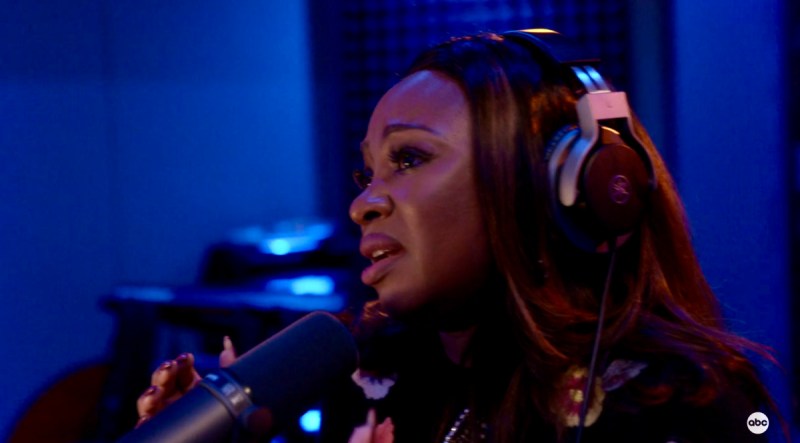 In a still from the television show "Queens" Naturi Naughton (Jill) raps into a microphone with oversized headphones against a blue background