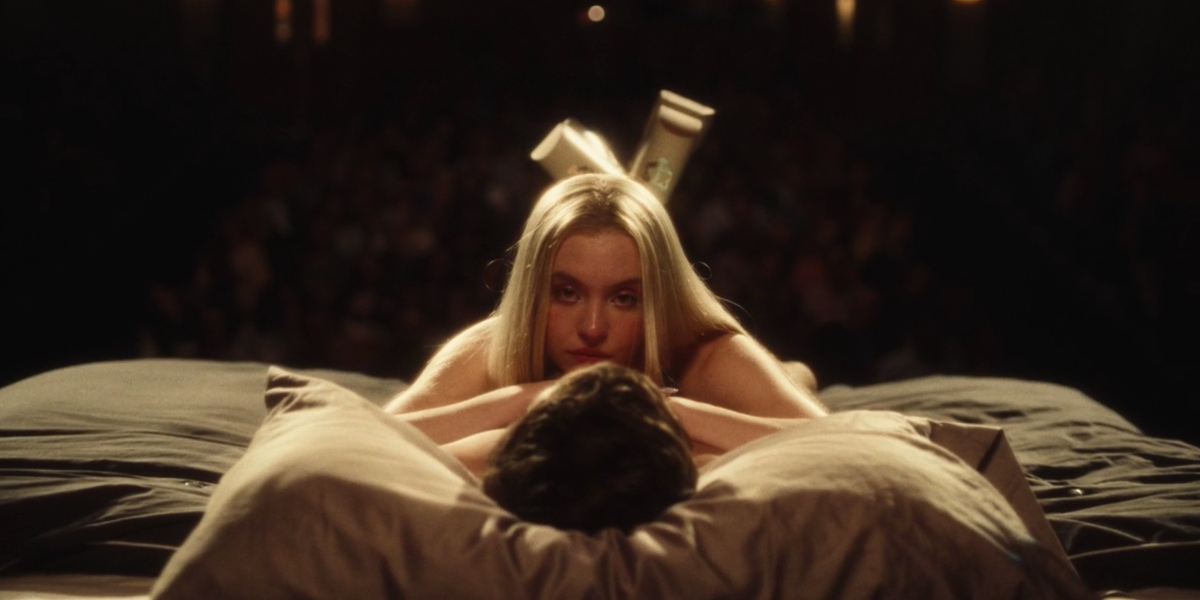 Cassie lies on top of Nate with her legs crossed behind her. She's looking toward the camera. Behind her we see an audience as if the bed is on stage.