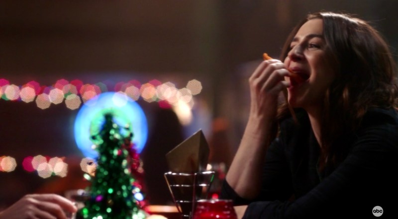 Amelia Shepherd smiles as she takes a big bite out of a sweet potato fry inside of Joe's Bar, with twinkly lights around her.