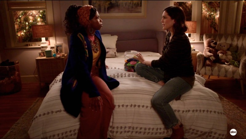 Denise, a black woman with long dreadlocks and saved sides on her hair, and Sarah, a white woman with a short brunette bob, are sitting together on a bed with a stripped comforter at night.