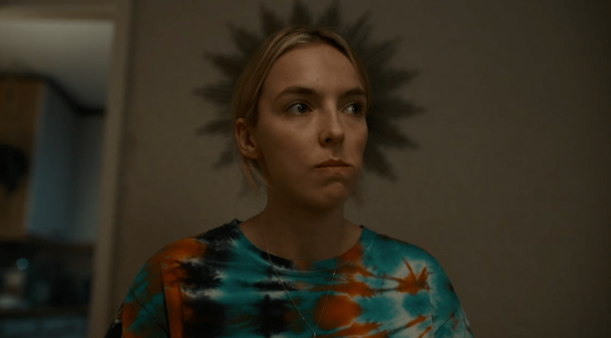 Villanelle (Jodie Comer) of Killing Eve wears a tie-dye shirt. A sunburst mirror on the wall behind her gives the appearance that she's wearing a crown.