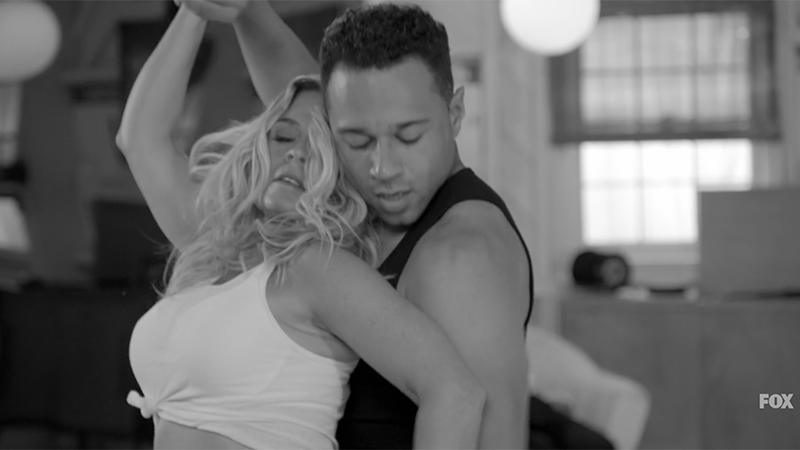 A black and white still of Cat in a white tank top and Corbin in a black tank top with Cat leaning back into Corbin with their hands above their head