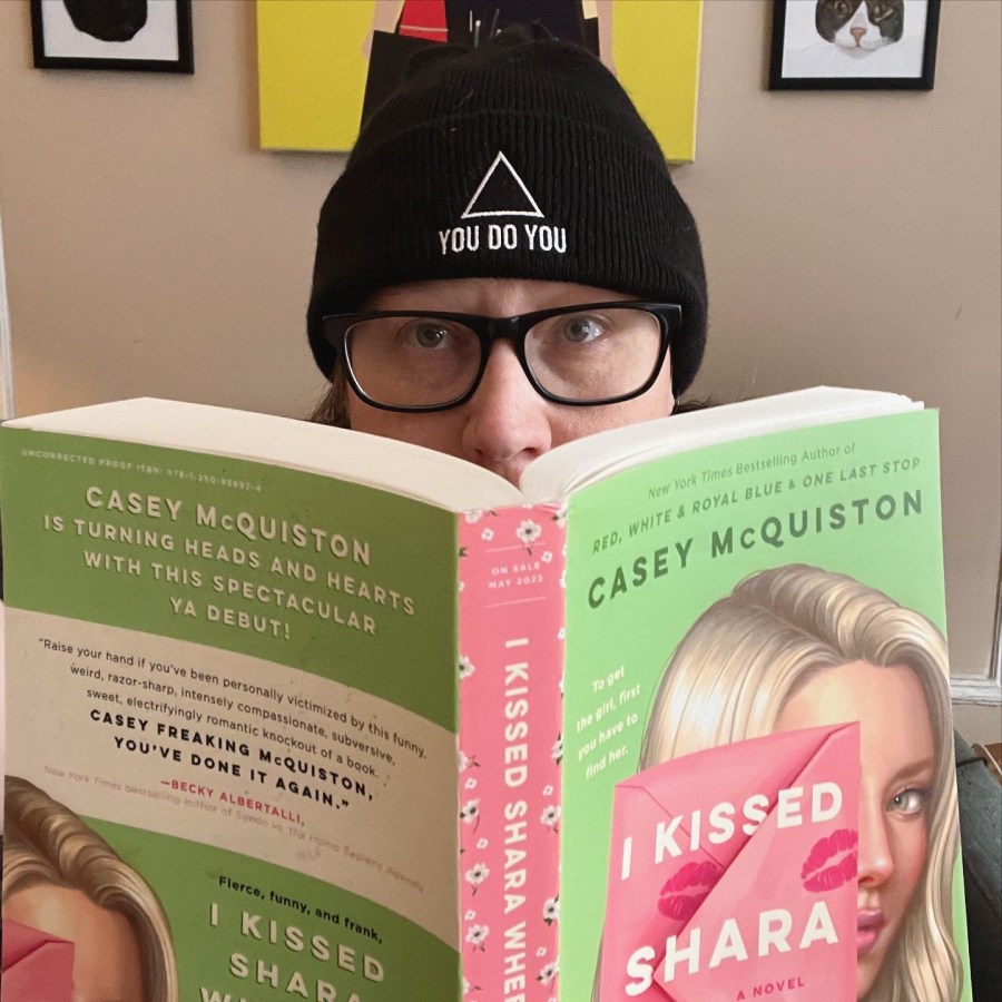 Heather peeks out from behind the book "I kissed Shara" by Casey McQuiston. Heather is a white woman wearing black glasses and one of our You Do You beanies.