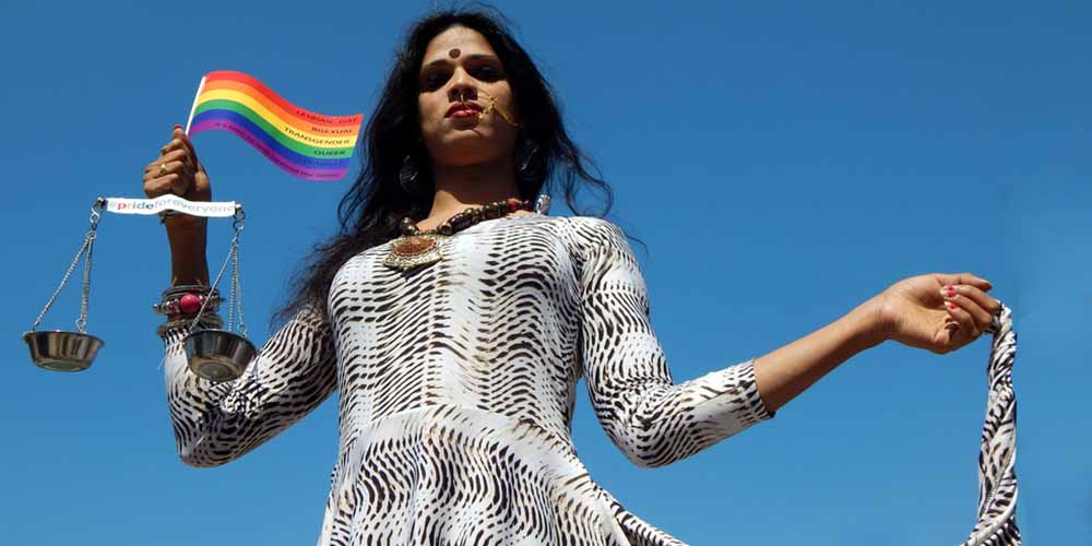 A feminine South Asian person in a black and white dress holds the hem of their dress in one hand and a Pride flag wit measuring scales on the other hand.