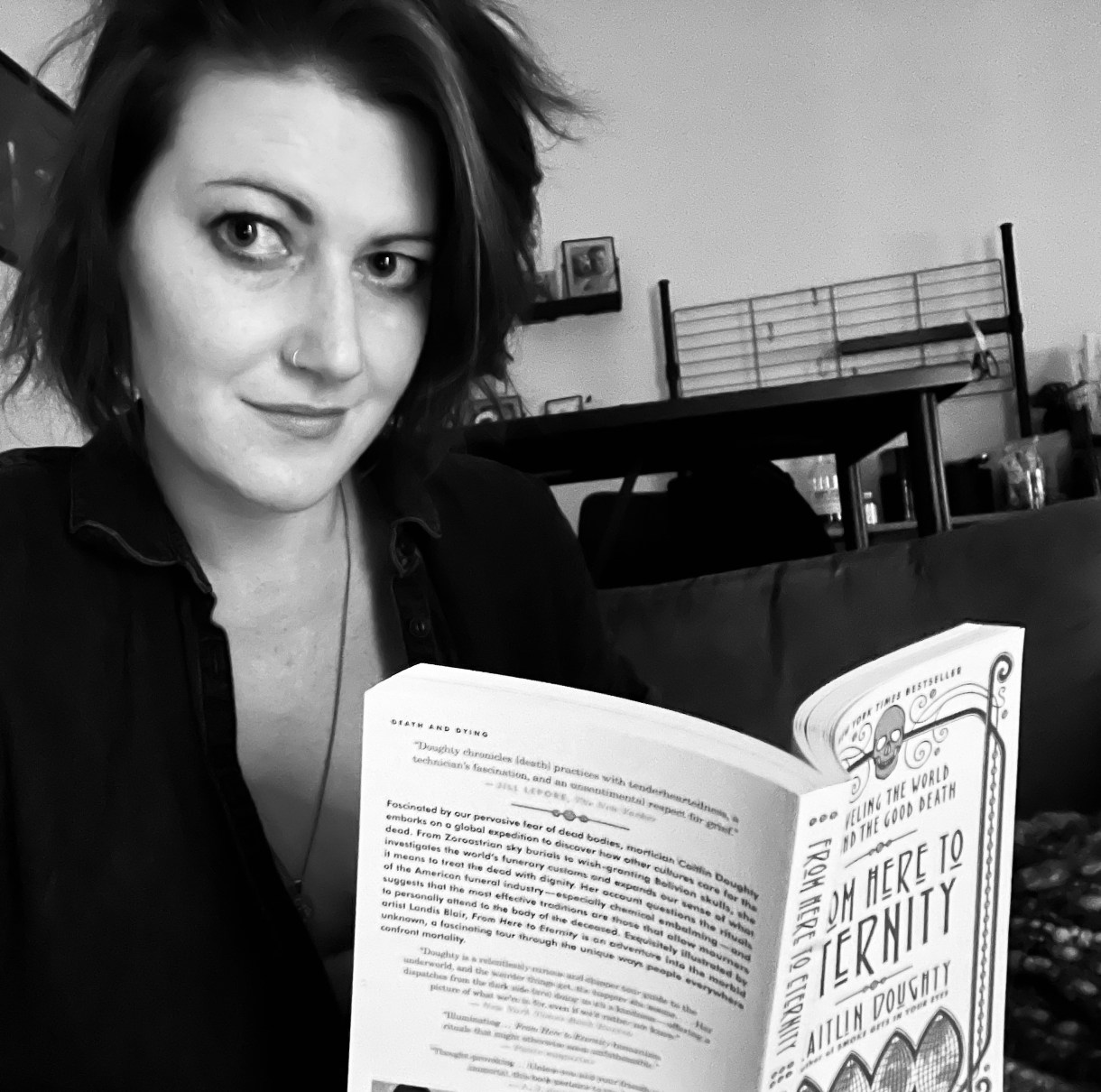 Meg reads "From Here to Eternity" but looks up and at the camera. Meg is a white woman who is wearing all black in this photo.