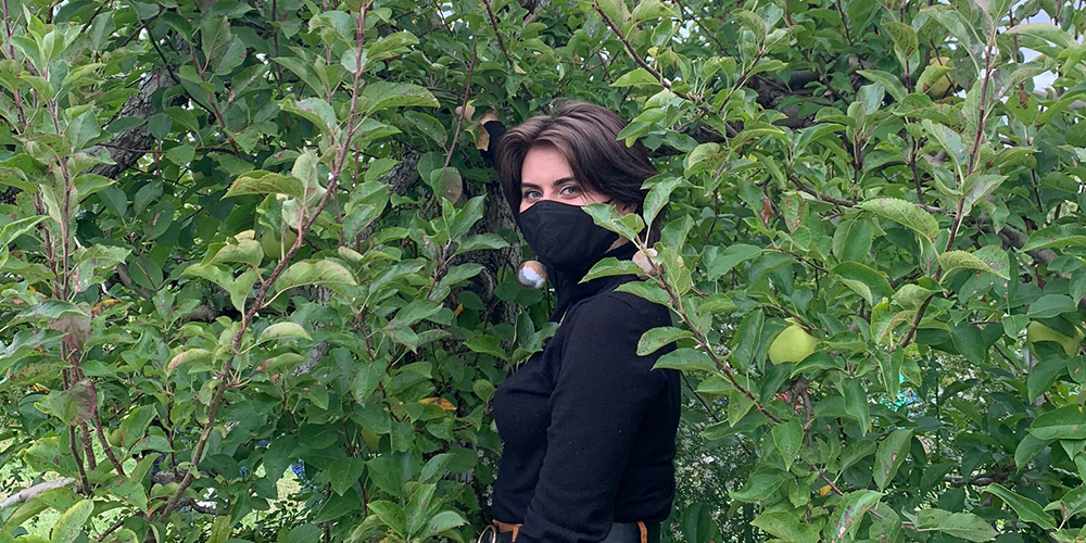 A fair-skinned person with a black long-sleeved top and black face mask stands amid green shrubbery.
