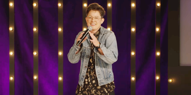 River Butcher in a jean jacket and black button-up standing on stage with a microphone laughing in front of a purple backdrop