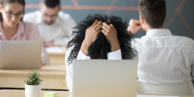 A frustrated Black woman feeling despair sits by her computer in a shared office holding her head in her hands.