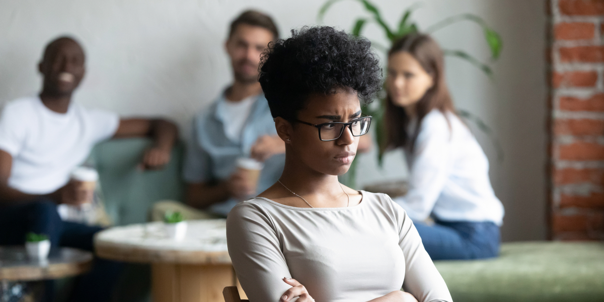 a woman sits in the foreground looking miserable and left out, while her girlfriend sits with two friends in the background, looking at her with dismay