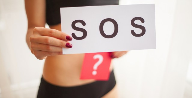 a white person in a black sports bra and briefs holds up a sign that says S O S and another sign that has a question mark on it, near their vulva/vaginal region