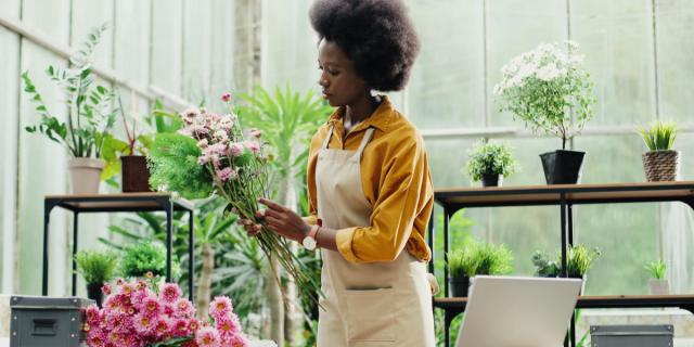 A Black woman arranges a bouquet of small pink flowers. She is wearing a mustard colored buttondown and a light brown apron. She is in a floral studio.