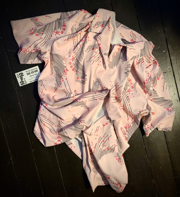 A polyester cool-toned pink short-sleeve button up top with an 80s or 90s looking black line and darker pink flower motif. The shirt is very loose and flowy which you can tell from the way it lies on the ground. There is a business card next to it for The 212 Club which is pronounced, according to the card, The Two Twelve Club. It's Amarillo address, 212 W. 6th ST. and phone number (806) 372-7997 are visible in the photo.