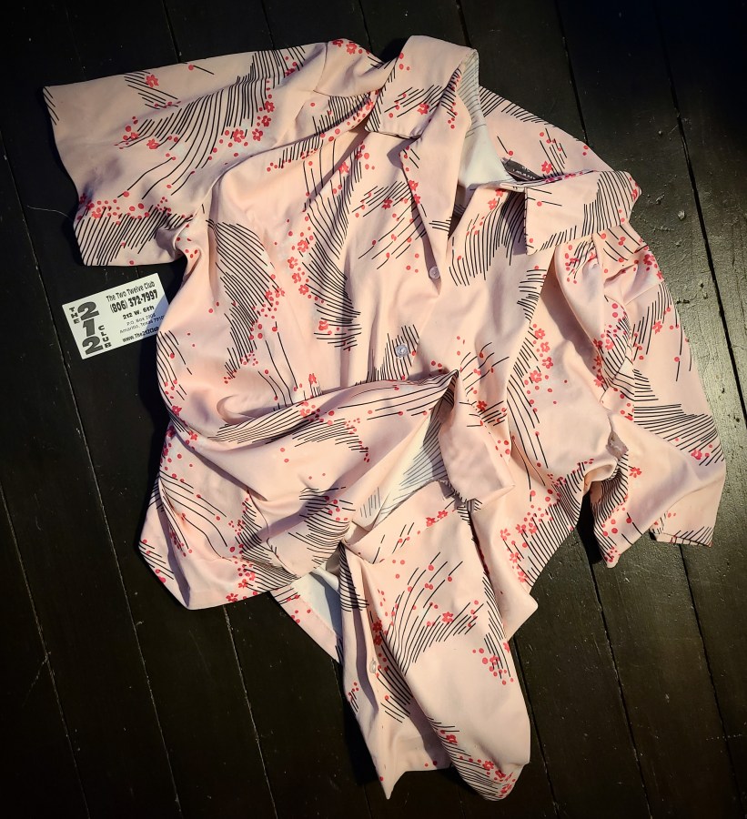 A polyester cool-toned pink short-sleeve button up top with an 80s or 90s looking black line and darker pink flower motif. The shirt is very loose and flowy which you can tell from the way it lies on the ground. There is a business card next to it for The 212 Club which is pronounced, according to the card, The Two Twelve Club. It's Amarillo address, 212 W. 6th ST. and phone number (806) 372-7997 are visible in the photo.