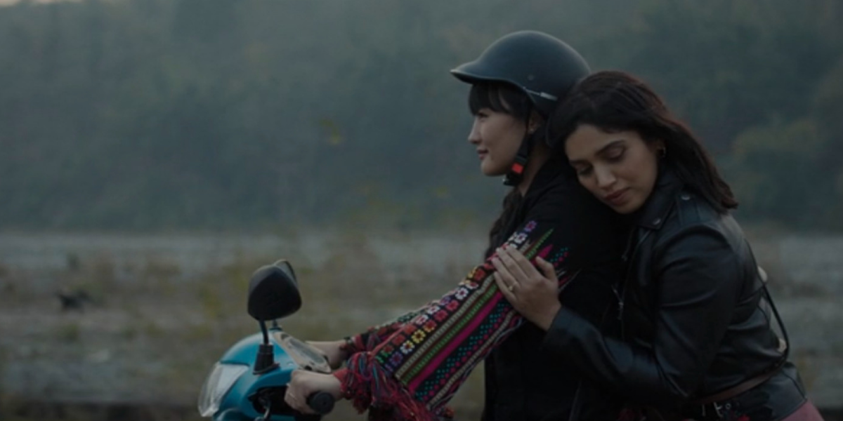 Two women ride a moped together, one is tenderly holding on with her eyes gently closed as the other drives.