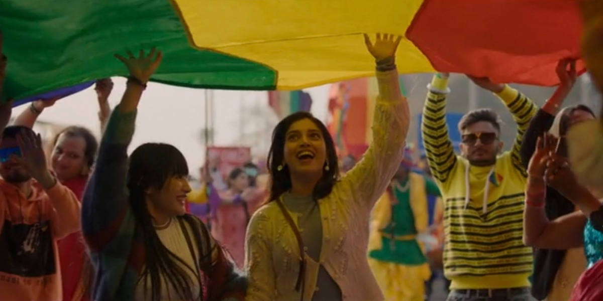 Two girls holding hands and smiling under a huge flag at a pride event in India