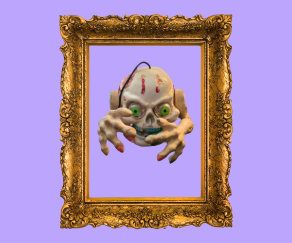 A halloween decoration skull with gummy arms coming out of the sides floats within a golden frame. It has green sinister eyes and a spooky grin.